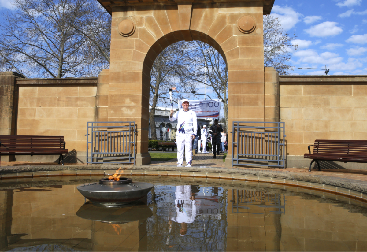 Wagga Mayor Councillor Dallas Tout walks under the Memorial Arch at Victory Memorial Gardens, with Eternal Flame in foreground and Defence personnel carrying the Legacy Centenary Torch Relay banner in the background